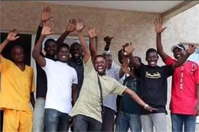 [ai] A group of eleven men standing together, smiling and raising their hands, in front of a building. They are wearing a mix of casual clothing in various colors.